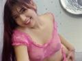 BrendaaPierce with long red hair, smiling, wearing a sheer pink top that reveals her small tits, and pink side-tie panties, leaning forward in a bathroom.