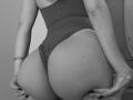 SugarrX's big, round butt in a tight, dark one-piece, holding it with both hands, showing off her ass and a silver bracelet on her wrist. Black nail polish.