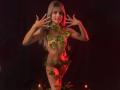 SheylaSpooner with green leafy vines covering her tits and pussy, standing naked with hands up, smiling, in a dark red-lit studio, surrounded by candles on the floor.