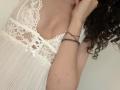 Os seios cobertos de renda de SweetClaraa, em um camisolo branco com pregas, com cabelos castanhos cacheados e uma pulseira vermelha no pulso direito, segurando o cabelo perto do rosto..