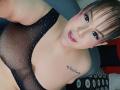 GiiannaMorrys with pink eyelids, black mesh top, small white dots, visible tattoo on right shoulder, looking at camera, smiling, brown hair, bangs, gray couch, pink lipstick.