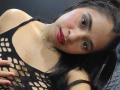 EmilyVidal with dark hair, red lipstick, and black fishnet top, showing her tits through the net, wearing a silver necklace, and looking at the camera with red nails.