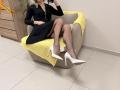 Rorria est assise sur une chaise beige avec une couverture jaune, vêtue d'une robe noire, de bas résille et de talons blancs, les jambes croisées, montrant ses fesses et ses talons blancs..