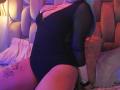 MaddieLesYoux in a black, sheer-armed, deep-v, one-piece, showing her tits, with blue hair, tattoo on her thigh, sitting on a bed, hands on her chest.