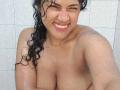 IvannaDuran with wet curly black hair, smiling, showing her large tits, and glistening skin in a shower, arms crossed, eyes squinted, pink lipstick, and a playful expression.