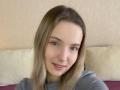 NicoleSpritzer has light blonde hair, green eyes, and a cute smile with pink lips. She's wearing a gray top, and her face is slightly turned to the right.