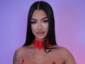 NatalieDelices with long black hair, red collar, and gold ring, topless, showing tattoos, with pink lipstick and red nails, looking at camera with a slight smile.