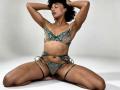 RoldanStefy with curly black hair, eyes closed, wearing green lace bra and panties, sitting with legs spread, arms behind head, hands in curly hair, showing tan skin and small tits.