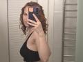 ChayaParrett with curly red hair, black sports bra, holding phone, showing sideboob, small ring on finger, watch on wrist, mirror selfie, towel rack in background.