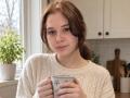 HoaGunby with brown hair in a ponytail, wearing a white knit sweater, holding a blue-patterned mug, standing in a kitchen, smiling softly. Her tits are small and she's cute.