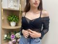 Rioria with black sheer off-shoulder top, showing her small tits, and blue jeans, holding her top, wearing gold hoop earrings, brown hair, and pink lipstick.