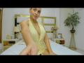 EvelynSwift with medium tits, wearing a yellow halter dress, sitting on a bed, smiling at the camera, right arm extended, in a bedroom with a gold headboard and plant.