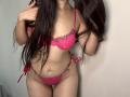 Tisia with long brown hair, wearing pink lace bra and panties, holding hair with right hand, showing her tits and pussy, standing against a plain wall.