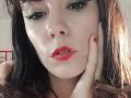 OrianaLaFrancaise with pale skin, dark brown hair, red lipstick, and blue eyeshadow, touching her cheek with red-manicured fingers, looking slightly to the side with parted lips.