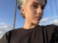 HanhFontillas with short blonde hair, brown eyes, black shirt, and earrings, looking serious, standing next to a metal chain, clear blue sky, and scattered clouds in the background.