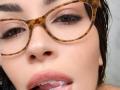 ZoeyMarie's face with tortoiseshell glasses, brown eyes, and pink lips, tongue slightly out with white fluid on her upper lip. Her dark wet hair is partially visible.