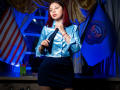 StephanieSun with red hair, blue shiny blouse, black skirt, holding black tie, looking confident, standing in front of American flag and Presidential seal. Her ass is cute.