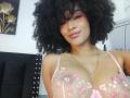 SnackTime with curly black hair, nose ring, and pink floral bra, showing her big tits, smiling at the camera, wearing a gold necklace.