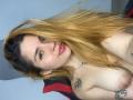 AbbyJanes with long blonde to brown ombre hair, nose ring, and pink lipstick, topless, showing her small tits and tattoos, smiling at the camera.