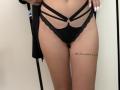 MysteryLaddy's black strappy lingerie with lace edges, showing a tattoo on her right thigh, a belly button piercing, and white nails, standing against a white wall.