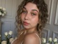 Sanella with curly brown hair, green eyes, and pink lips, wearing a white top, surrounded by white roses, showing her bare shoulders and subtle cleavage.