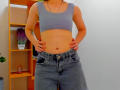 Steisii with a flat stomach, wearing a gray sports bra and high-waisted blue jeans, hands on hips, showing a tattoo on her left arm, and a necklace.