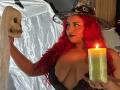 Estefy69 with red curly hair, witch hat, and black lace dress, holding skull and candle, showing big tits, looking at skull, candle lit, warm light, sexy, witchy vibe.