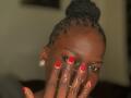 Close-up of KharlaQueen with dark skin and short, twisted hair in a bun. She covers her face with her hand, showing red-painted nails with white heart and line designs. She wears small, clear earrings. Her eyes are slightly open, looking to the side. The background is blurred. The image has a warm, soft light. Her eyebrows are slightly raised. Her nails are shiny and well-manicured.