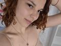 ChayaParrett with curly red hair, pale skin, and subtle smile, wearing a beige top and necklace, looking at camera with slightly raised arm. Boobs visible, small to medium.