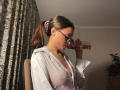 KassandraDalal with glasses, white shirt, visible nipples, brown ponytail, drinking from a pink bottle, seated, looking down, in a dimly lit room.