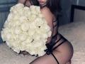 MilagrosEdmisten kneels on a white furry surface, holding a large bouquet of white roses, wearing black lace lingerie. Her curvy body and sexy pose create intense sexual tension.