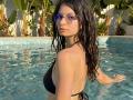 VikkiRi with wet black bikini, small tits, blue-tinted sunglasses, and long dark hair in a sunny pool, looking over her shoulder. Her hand rests in the water.