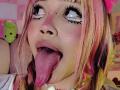 LiMiuu with pink and blonde hair, big black eyeliner eyes looking up, tongue out, and lip piercing, showing her pink tongue and tongue piercing.