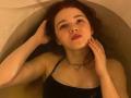 KatiRokisky, with red hair, in a bathtub, wearing a black halter top, hands in water, looking up, slightly parted red lips, necklace with a small ring.