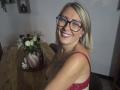 AmandalaFrancaise with blonde hair, glasses, and red lace bra, smiling at the camera, showing her tits and tattoo, standing by a wooden table with a floral vase.