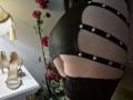 Rouzis Hintern in schwarzem Leder mit Metallriemen, der einen wohlgeformten Po zeigt, trägt schwarze Spitzenstrümpfe, dazu beige High Heels und eine Wand mit roten Blumen..