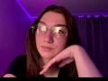 SunnyKlusman with glasses, nose ring, and pouty lips, resting chin on hand, purple lighting, brown hair, wearing black shirt, reflective glasses, medium skin tone, soft expression.