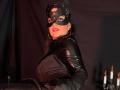 TamaraFoxy, avec un masque en cuir noir, une tenue brillante en latex noir et des gants, tenant un fouet en cuir noir. Elle a de grandes seins, des boucles d'oreilles en forme de cerceaux et du rouge à lèvres, dans un éclairage tamisé..