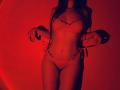 Tisia with red-lit skin wearing lace lingerie, holding strings of her thong, showing her tits and pussy, standing with long dark hair, no face visible.