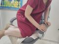 JoannaSia in a red dress, kneeling on a gray chair, white socks, hands on the seat, showing her pale ass and small tits, in a medical or clinic setting.