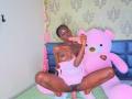KharlaQueen, dark-skinned, topless with small tits, sits on bed with pink stuffed bear, sucking pink dildo, legs spread, visible pussy, in pink and green room.