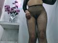 EmhilyDiaz trägt schwarze Netzstrumpfhosen, durch die ihre Vulva durch das Netz hindurch sichtbar ist. Ihre Hand ruht auf ihrem Oberschenkel. Lila Blumen in einer Vase stehen auf einem weißen Regal..