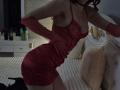 Rozalin in a red, sheer, lace-up dress, showing off her tits and ass, wearing long red gloves, black high heels, leaning forward in a dimly lit bedroom.