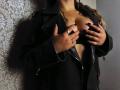 Niferttiti in a black leather jacket, hands covering her tits with black nails, gold rings, and a gold necklace, against a textured wall.