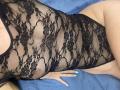 MoonLiza wearing a black, sheer lace bodysuit, exposing her tits through the floral pattern. Her hand with mint green nails rests on a blue surface.
