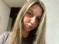 Sanella has long, straight blonde hair, light skin, and green eyes. She's wearing a brown ribbed shirt, with a neutral expression and slightly parted lips.