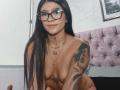 LuciadelaVega with long black hair, glasses, and tattoos, naked, showing small tits and a star tattoo on her chest, sitting on a bed with a pink tufted headboard.