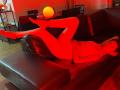 LilaaSalmon is lying on a black leather couch, wearing red sheer tights and a white top, with her legs up and arms resting on the couch. Red glow.