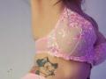 Close-up of ElixirShanna's upper body, showing a pink lace bra with floral embroidery, partially revealing her large, rounded breasts. Her left arm is raised, and a black and white tattoo of a smiling cat with a pink bow is visible on her right side. The image has a soft purple hue, and her skin is light. The texture of the lace is delicate and intricate.