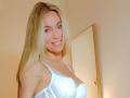 EllisaWoman with blonde hair, white lace bra, smiling, showing her tits, in a room with a door in the background. She's attractive, has light skin, and a cute face.
